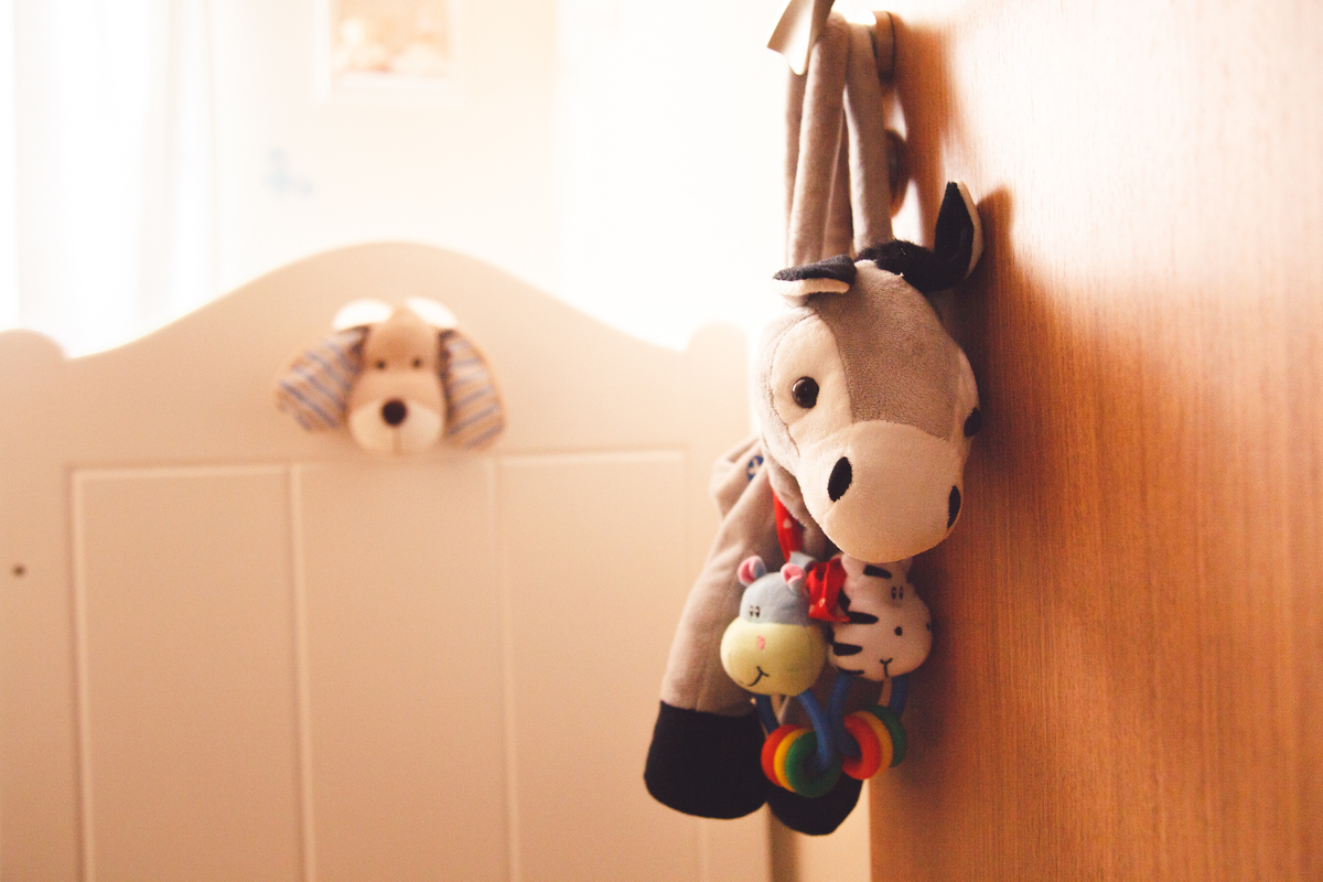 Detalhe de um burrinho de pelúcia pendurado na maçaneta da porta com o berço ao fundo, com a cabeça de um cachorrinho de pelúcia passando por um buraco em forma de coração na face do berço.