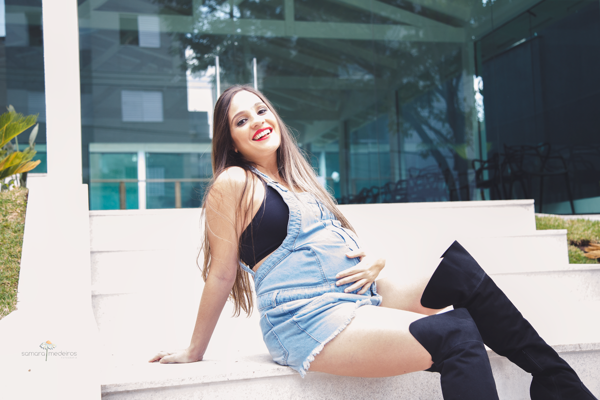 Mãe gestante sentada em uma escadaria branca, olhando para a foto e sorrindo, com uma mão na barriga e a outra apoiada no chão para sustentar o corpo.