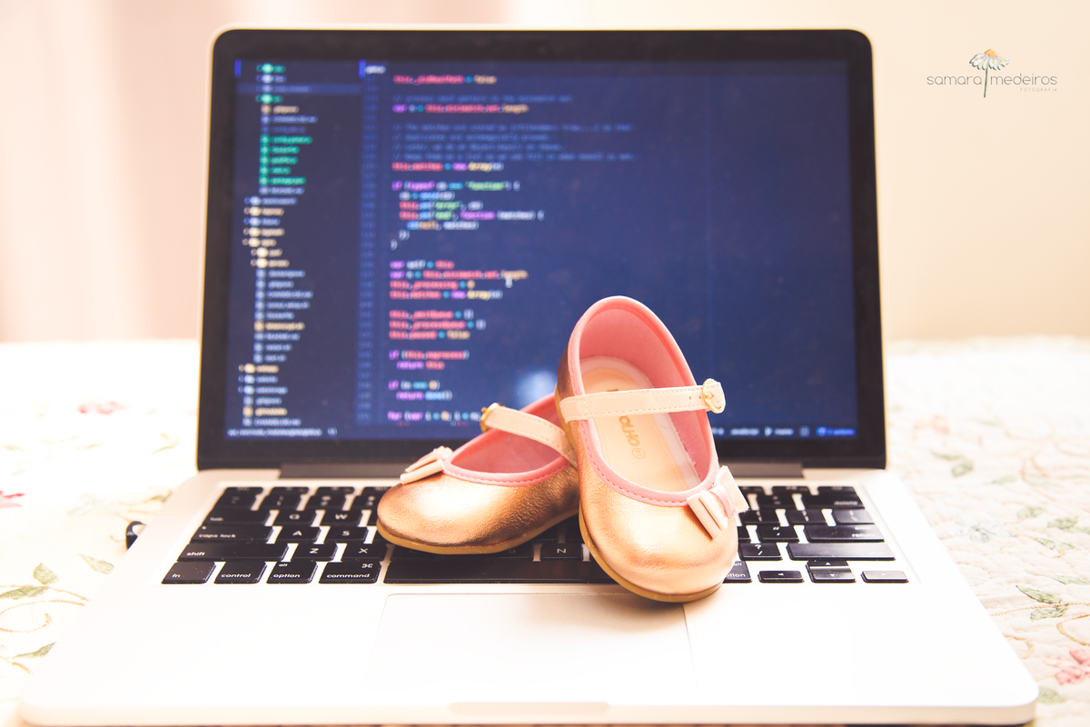 Detalhe de um computador com alguns códigos de programação na tela e um sapatinho de menina em cima do teclado, mostrando a união de duas partes da vida do pai.
