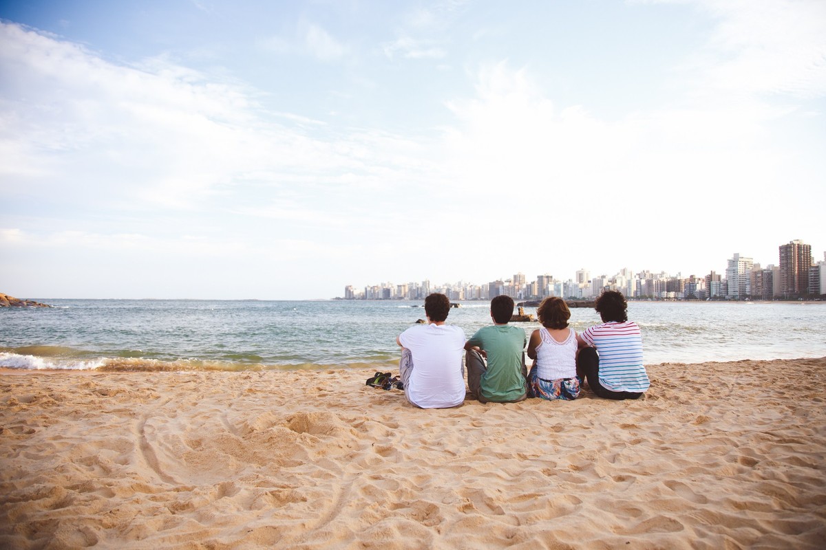 Três filhos adultos e sua mãe sentados na areia, na praia, de costas para a foto, olhando a cidade de Vitória.