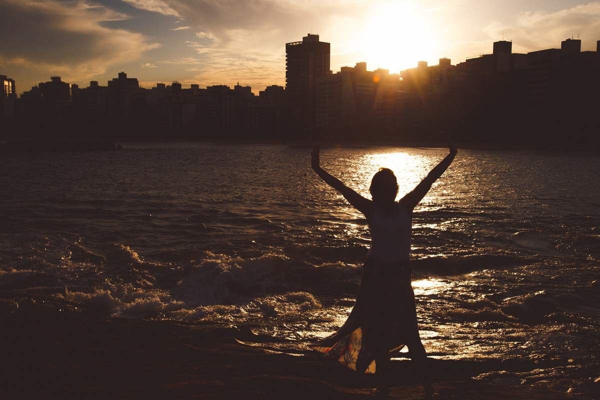Por-do-sol atrás da cidade de Vitória, refletindo no mar e a silhueta de uma mulher com os braços abertos, aproveitando o momento.