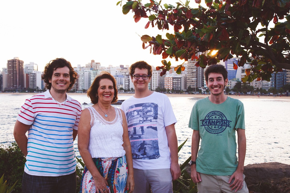 Retrato da família, mãe e três filhos adultos, todos sorrindo e olhando para a câmera, com o mar e a cidade de Vitória ao fundo.