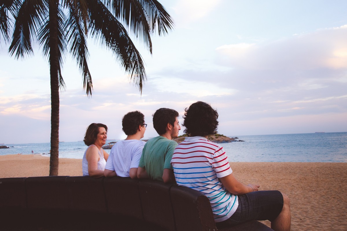 Família de mãe e três filhos adultos sentados em um banco na beira da praia, observando o por-do-sol.