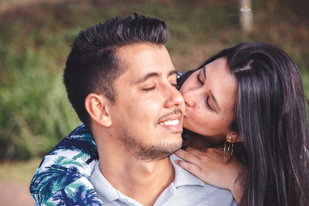 Moça abraçando o namorado pelas costas e dando um beijo em sua bochecha, ele de olhos fechados e sorrindo.
