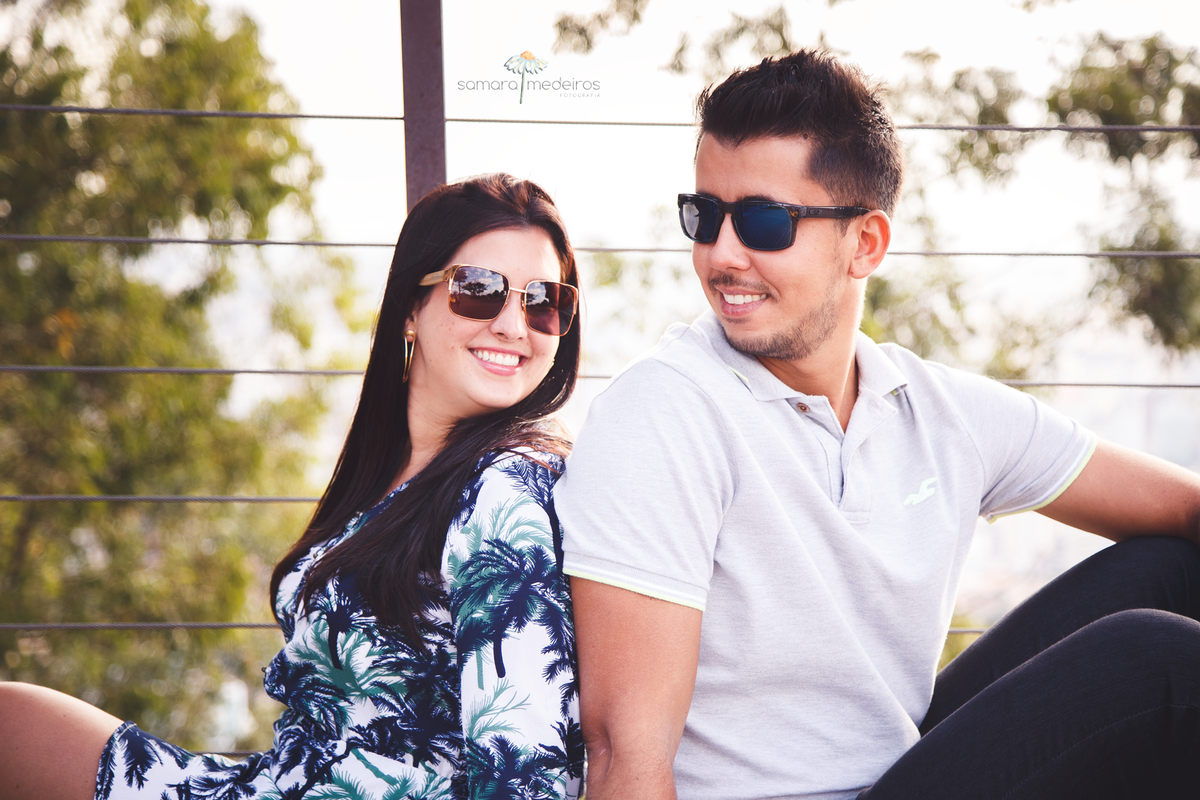 Casal sentado no chão de madeira, um de costas para o outro, se olhando meio de lado e sorrindo.