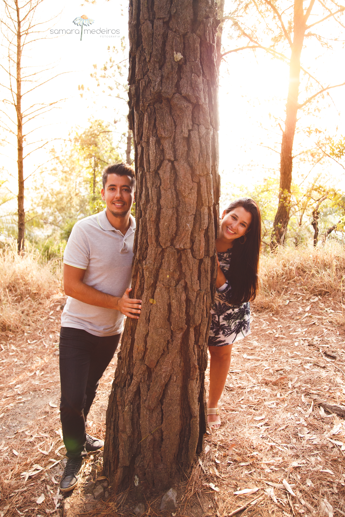 Casal posando para um ensaio fotográfico, brincando atrás de uma árvore e sorrindo, com o sol ao fundo.