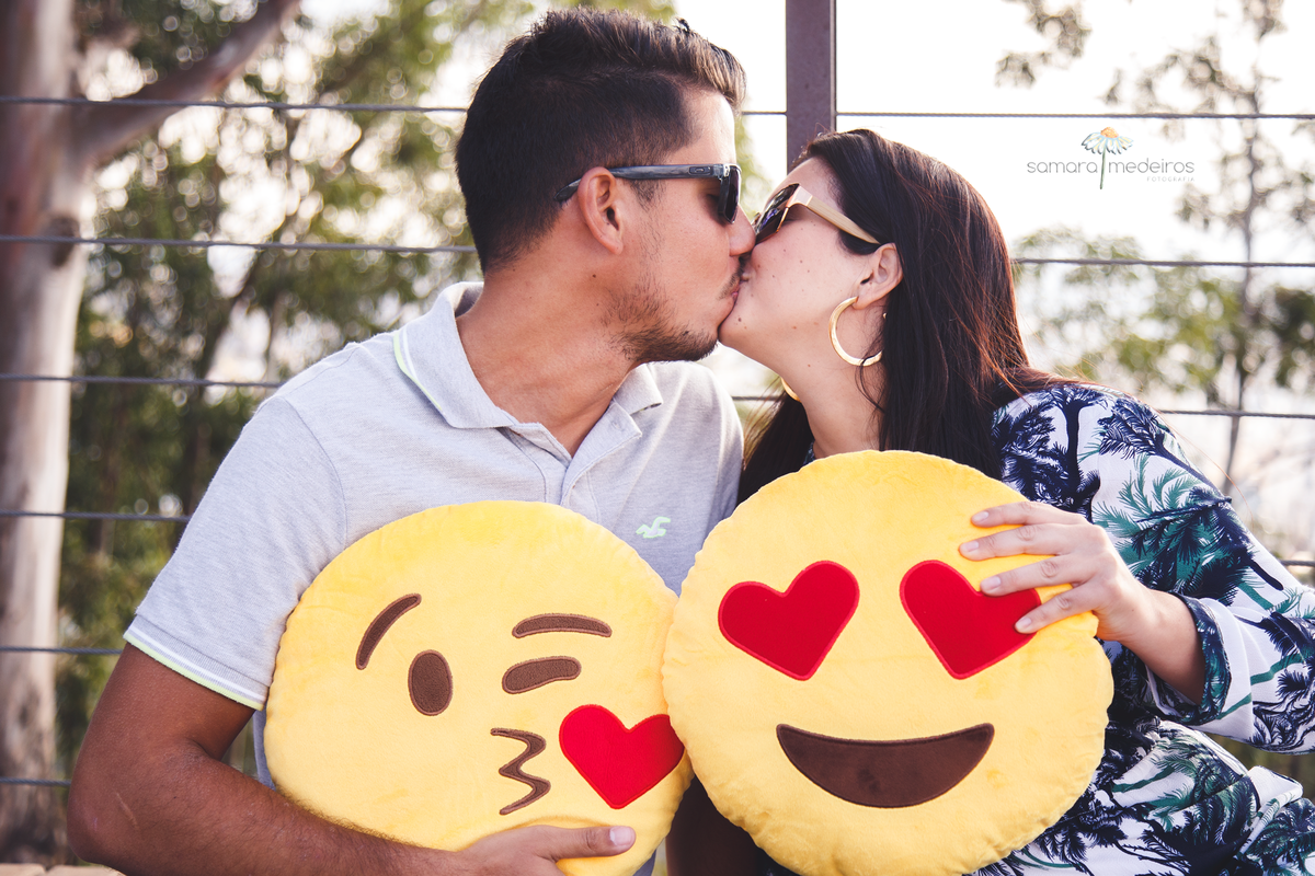 Casal sentado em um chão de madeira dando um beijo e segurando duas almofadas de emoticom.