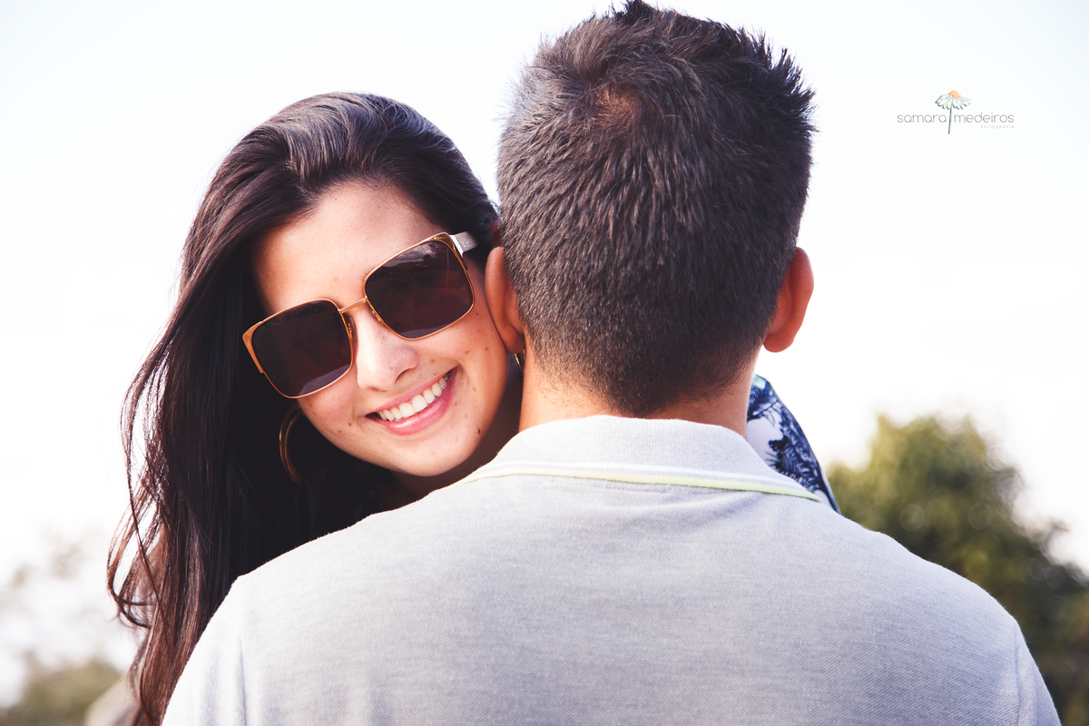 Casal abraçado, sendo o homem de costas e a moça de frente para câmera e sorrindo.