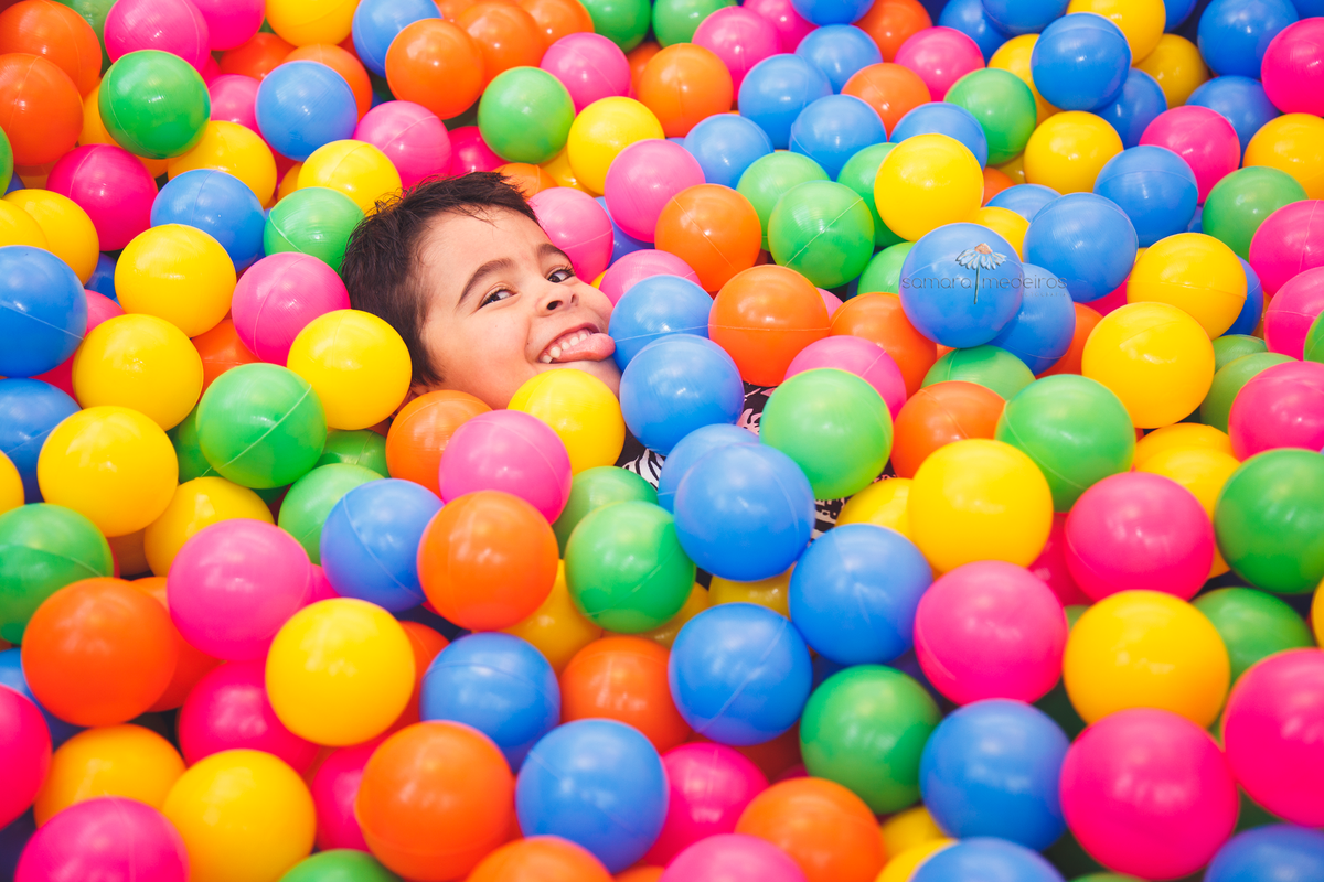 Mar de bolinhas coloridas com o rosto de uma criança semi afundado nas bolinhas, e fazendo uma careta.