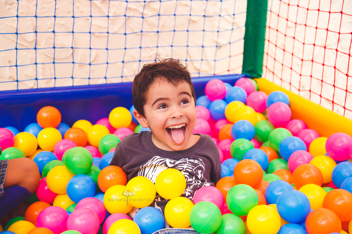 Criança em uma piscina de bolinhas sorrindo e fazendo careta para uma pessoa que não aparece na foto.