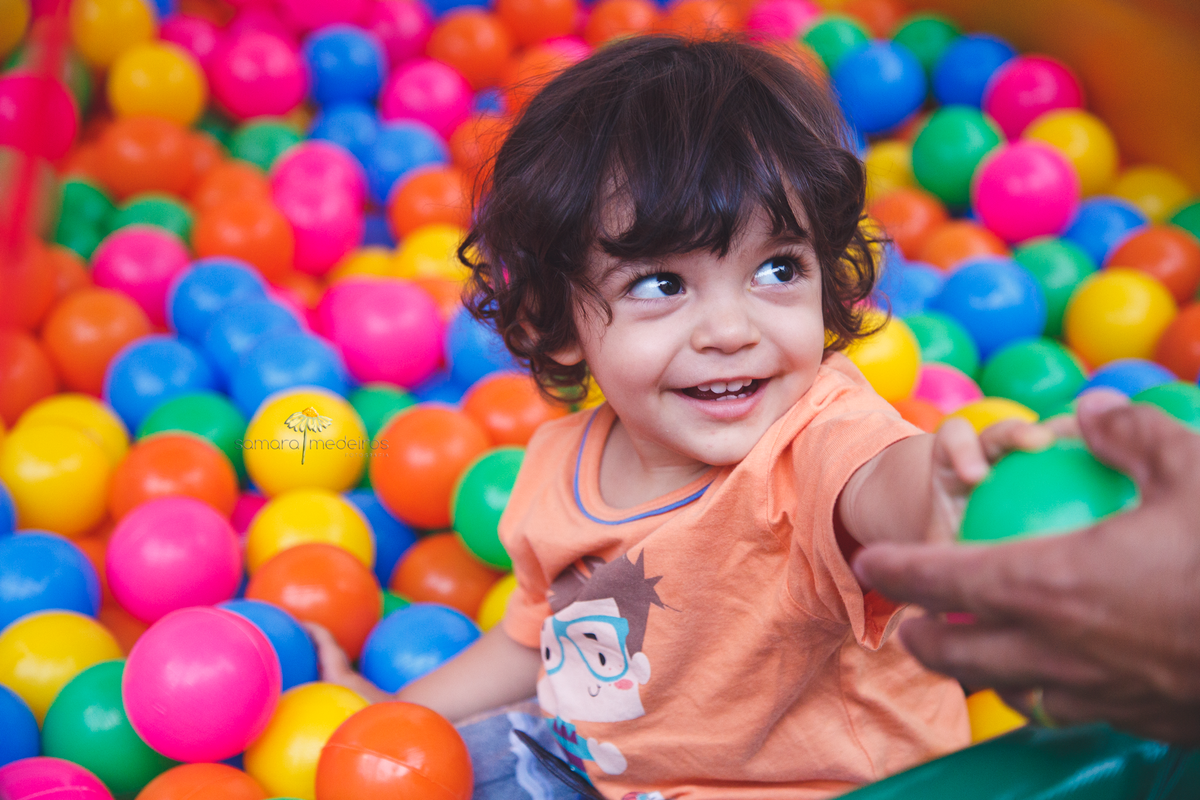 Criança pequena brincando na piscina de bolinha, sorrindo para seu pai.