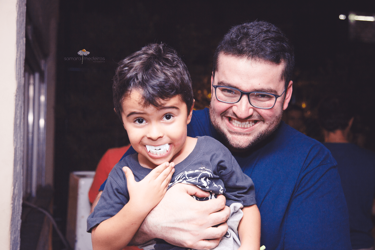 Criança no colo do padrinho com uma dentadura de vampiro na boca enquanto o padrinho também brinca mostrando os dentes.
