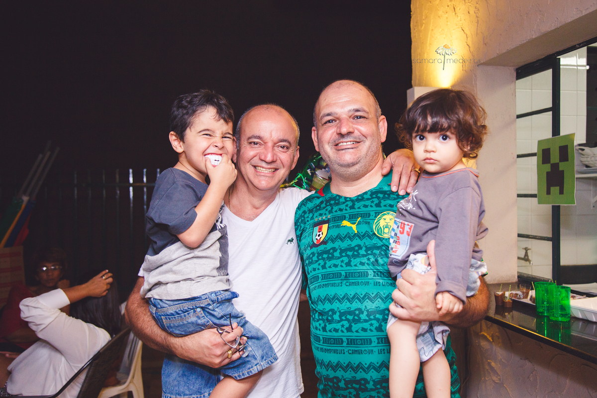 Dois adultos cada um com seu filho no colo, posando para foto e sorrindo, sendo que uma das crianças, o aniversariante, está fazendo gracinha com uma dentadura de vampiro.