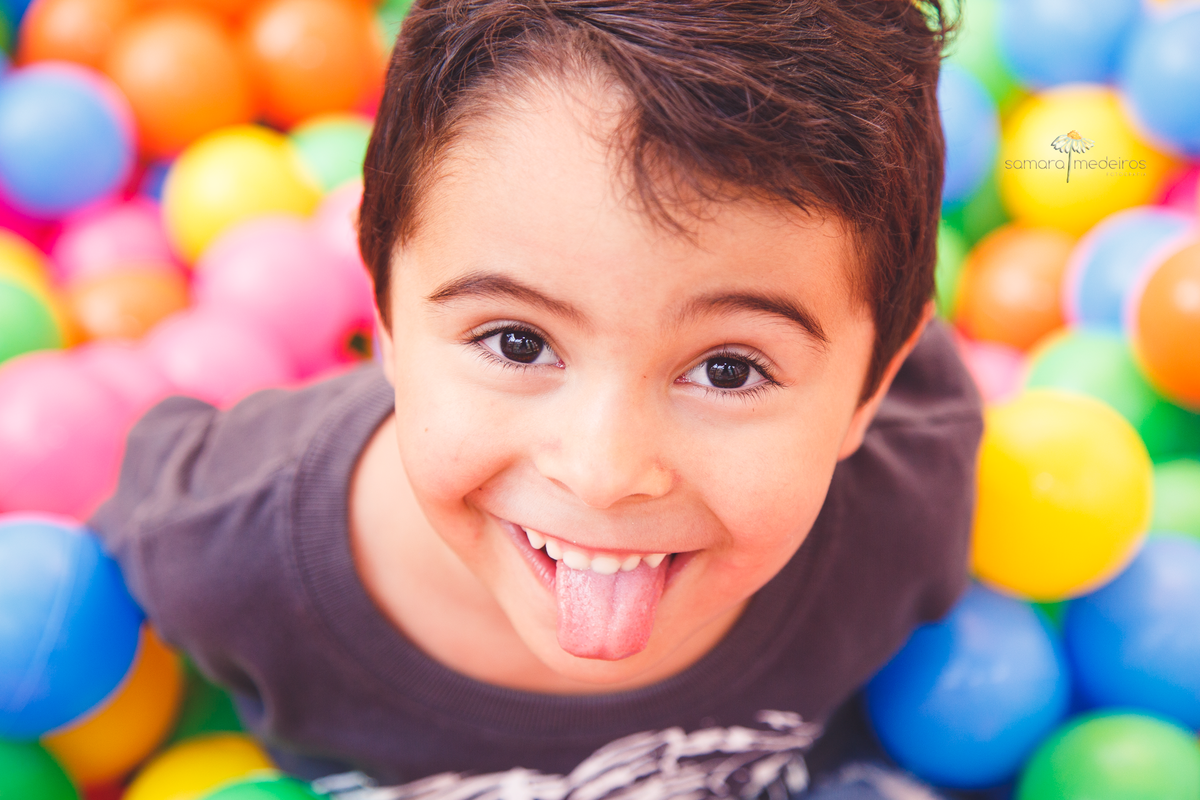 Close no rosto de uma criança, que sorri e faz língua para fotógrafa, sorrindo. Bolinhas coloridas de uma piscina de bolinhas ao fundo.