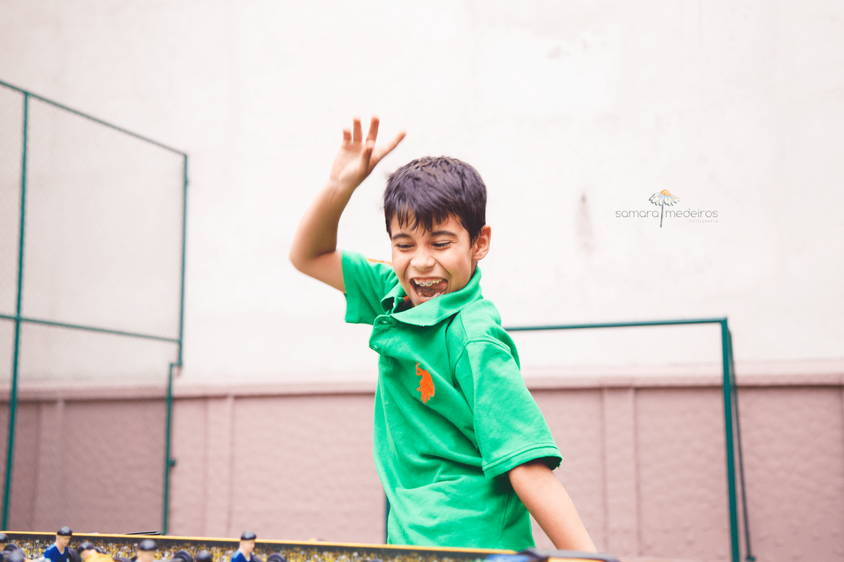 Foto espontânea de uma criança de 8 anos comemorando um gol no totó, sorrindo e com o braço para cima.