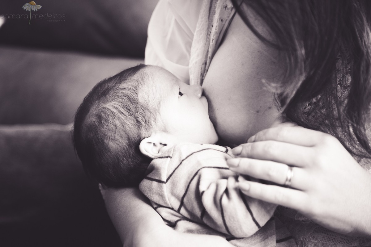 Foto em preto e branco do bebê mamando, com os olhos abertos, observando a mãe, que segura sua mãozinha.
