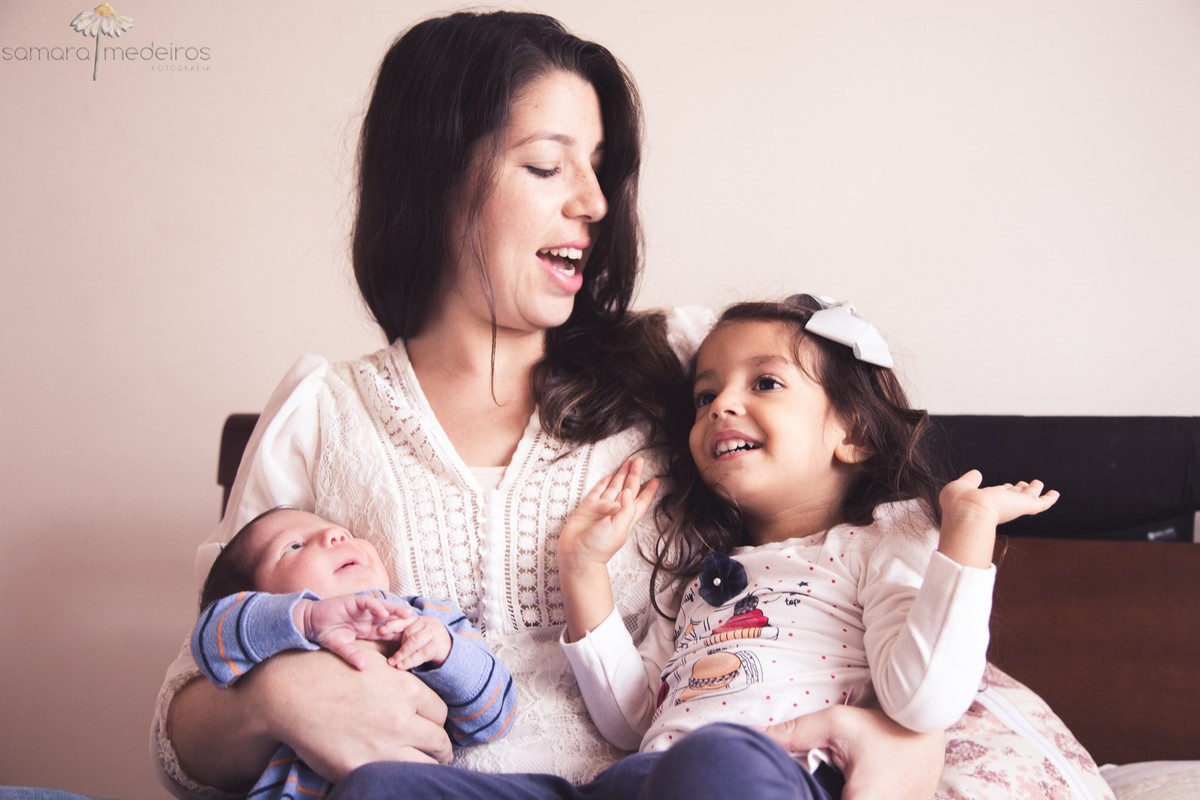 Mãe segurando os dois filhos, sendo um recém nascido de 15 dias, enquanto conversa e brinca com a filha mais velha, em sua casa em Belo Horizonte.