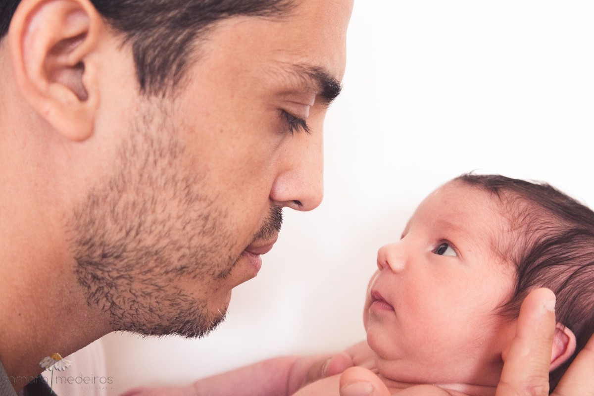 Pai segurando o bebê recém-nascido e os dois se olhando, durante sessão de fotos.