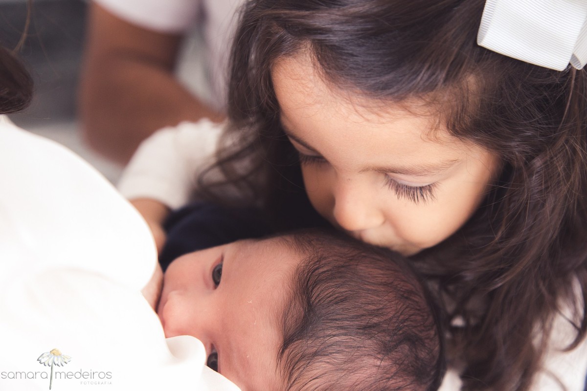 Irmã mais velha dá um beijo na cabecinha do bebê recém nascido enquanto ele mama.