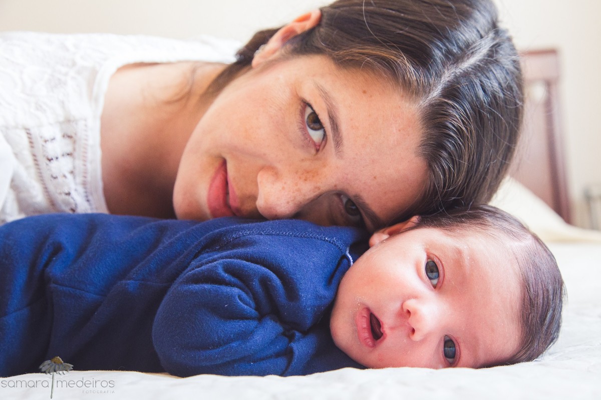 Bebê recém nascido deitado na cama, olhando para a foto e sua mãe com o rosto apoiado em suas costas, enquanto ela também olha para a foto.