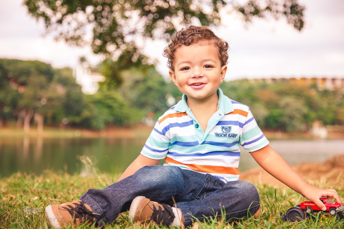 Criança sentada na grama brincando com um carrinho, olhando para a fotógrafa e sorrindo.