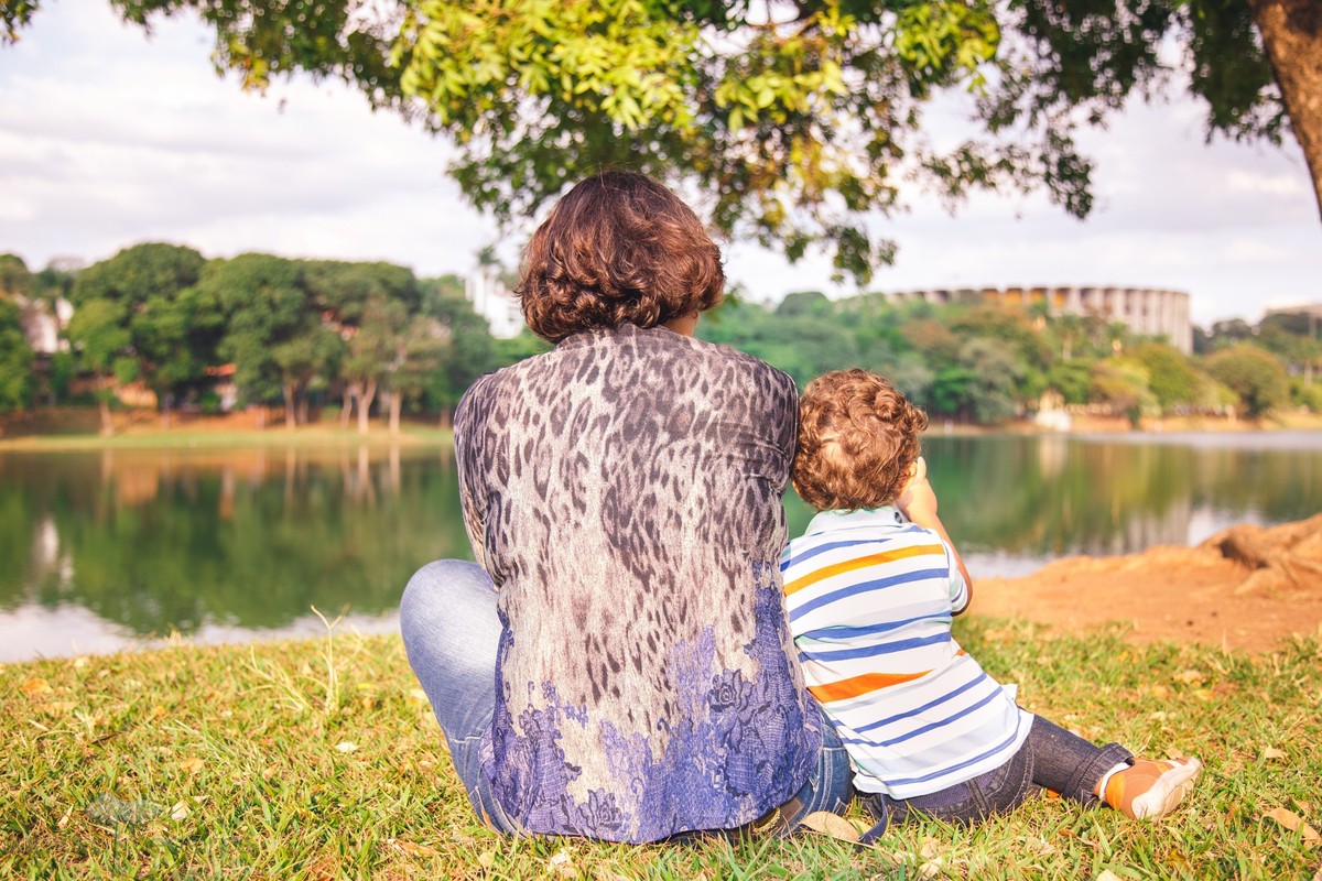 Mãe e filho lado a lado, de costas para a foto sentados na grama olhando para o Mineirão, ponto turístico de Belo Horizonte.
