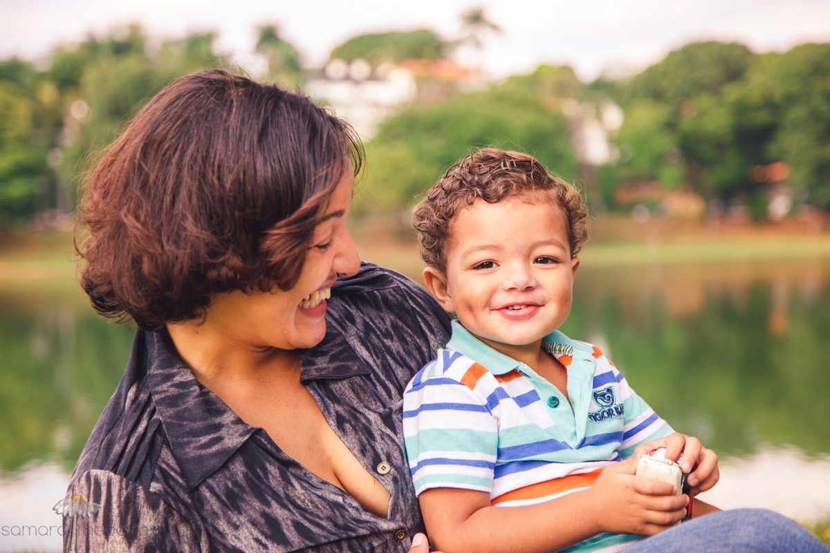 Mãe e filho sentados na grama brincando, sorrindo e se divertindo durante ensaio fotográfico de família.