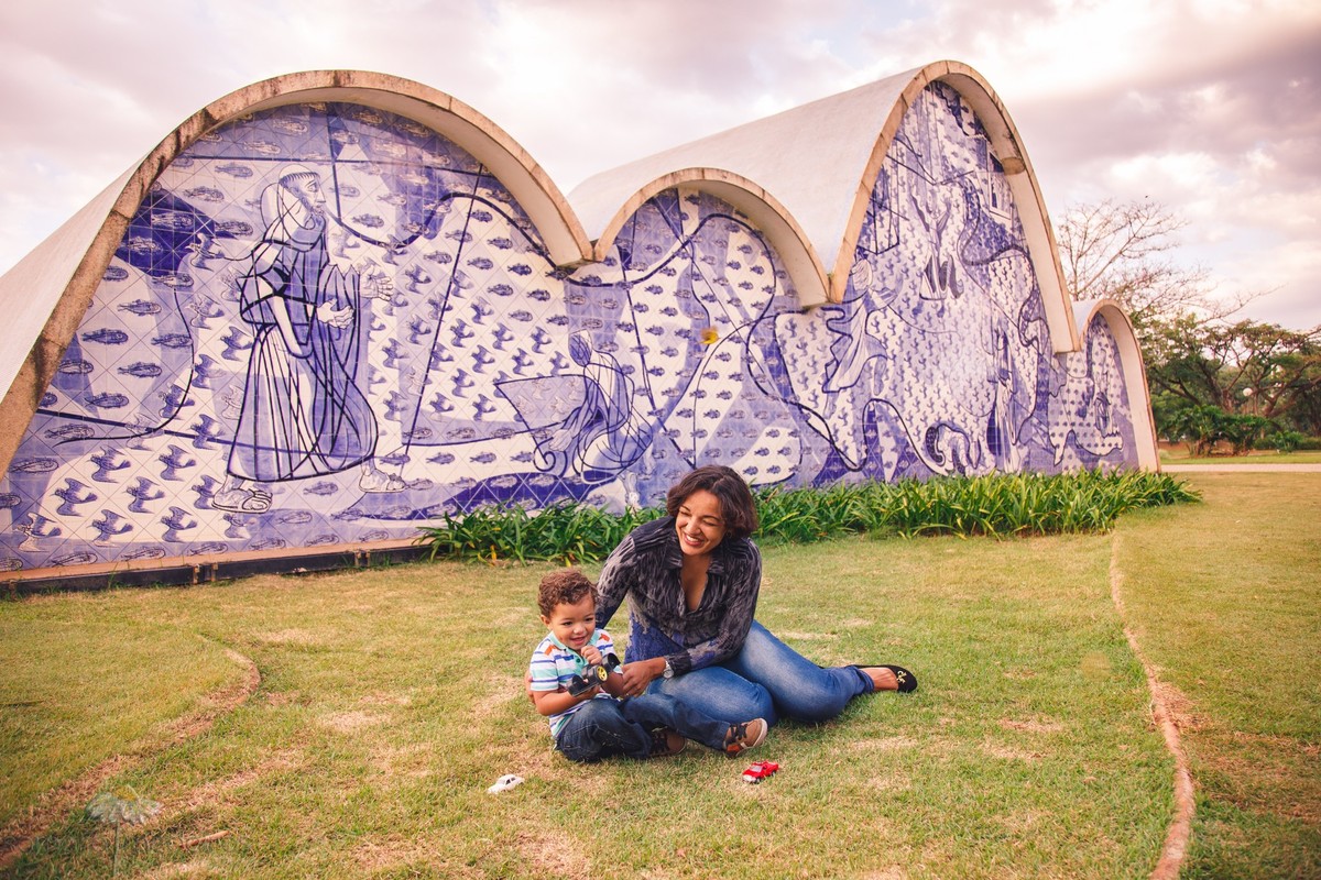Mãe e filho sentados na grama em frente à Igrejinha da Pampulha, brincando durante uma sessão fotográfica de família.
