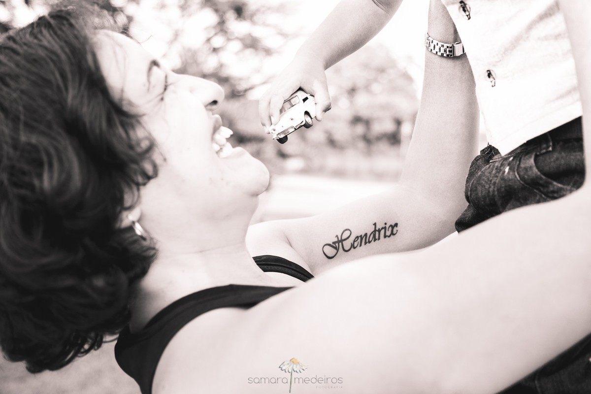 Foto em preto e branco de uma mãe carregando seu filho no alto, enquanto o foco fica na tatuagem do nome do filho escrito no braço da mãe.