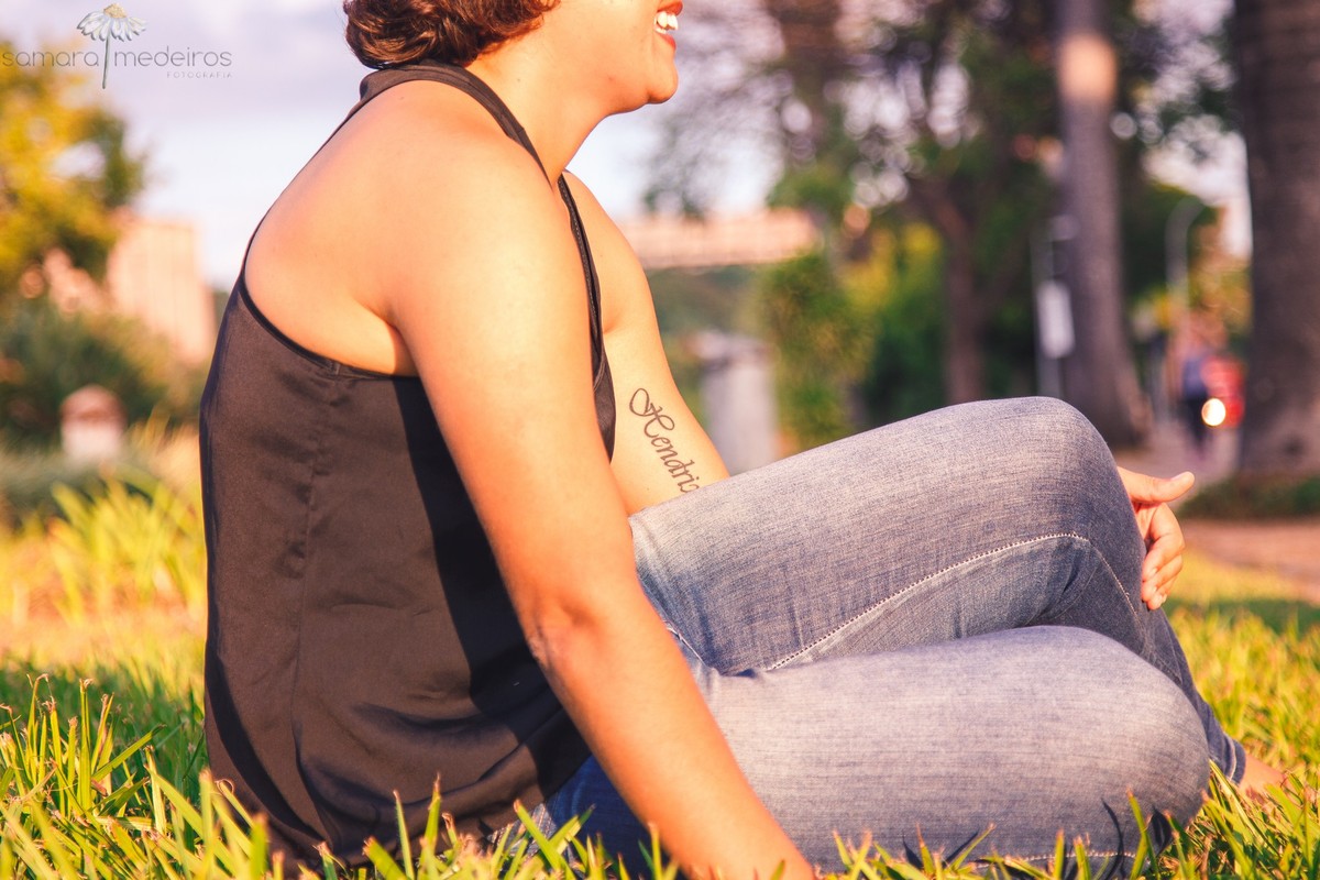 Mãe sentada na grama enquanto olha para o filho que está distante brincando. O foco da foto é a tatuagem com o nome da criança na parte interna do braço da mãe.