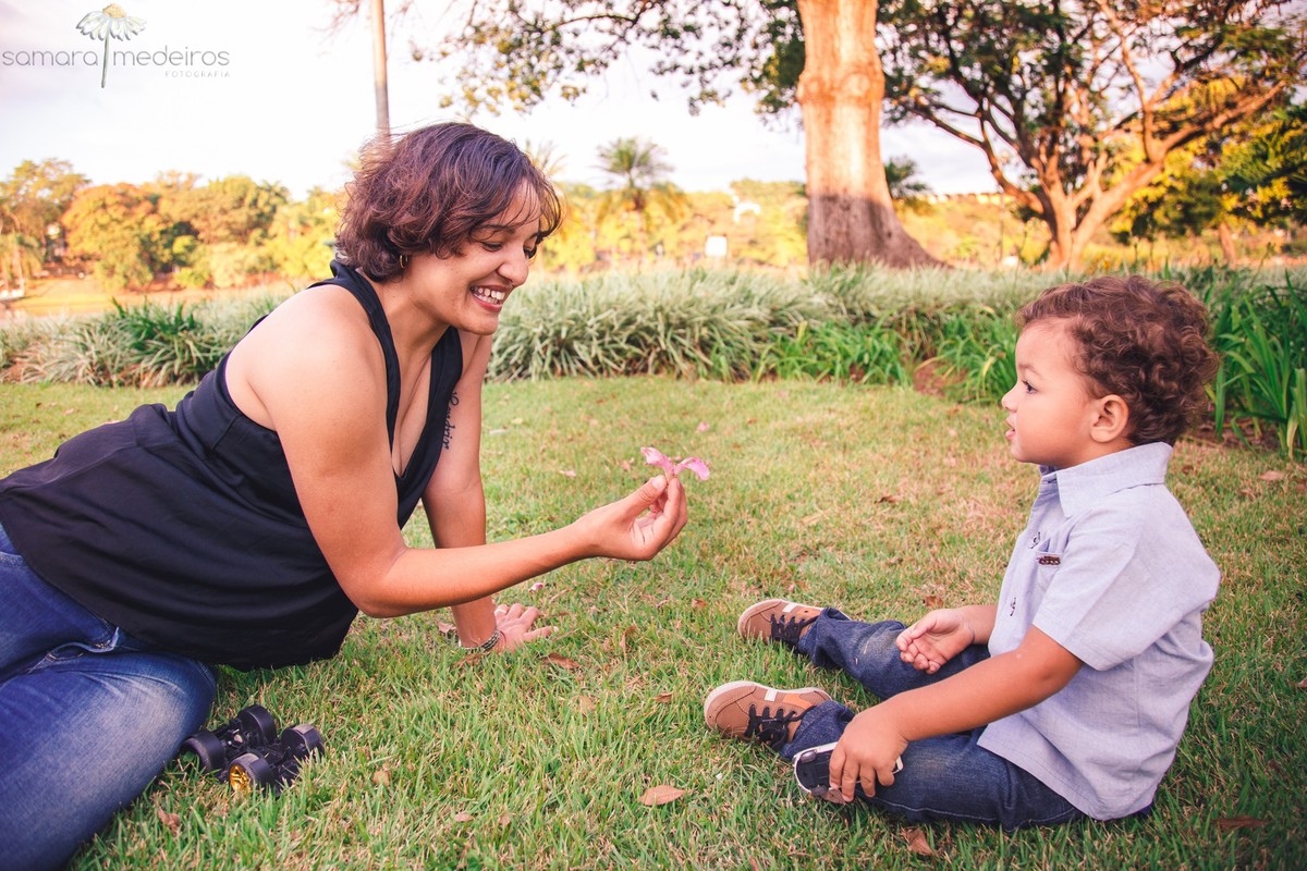 Mãe e filho sentados na grama enquanto a mãe oferece uma flor para o filho, enquanto posam para uma sessão de fotos de família.