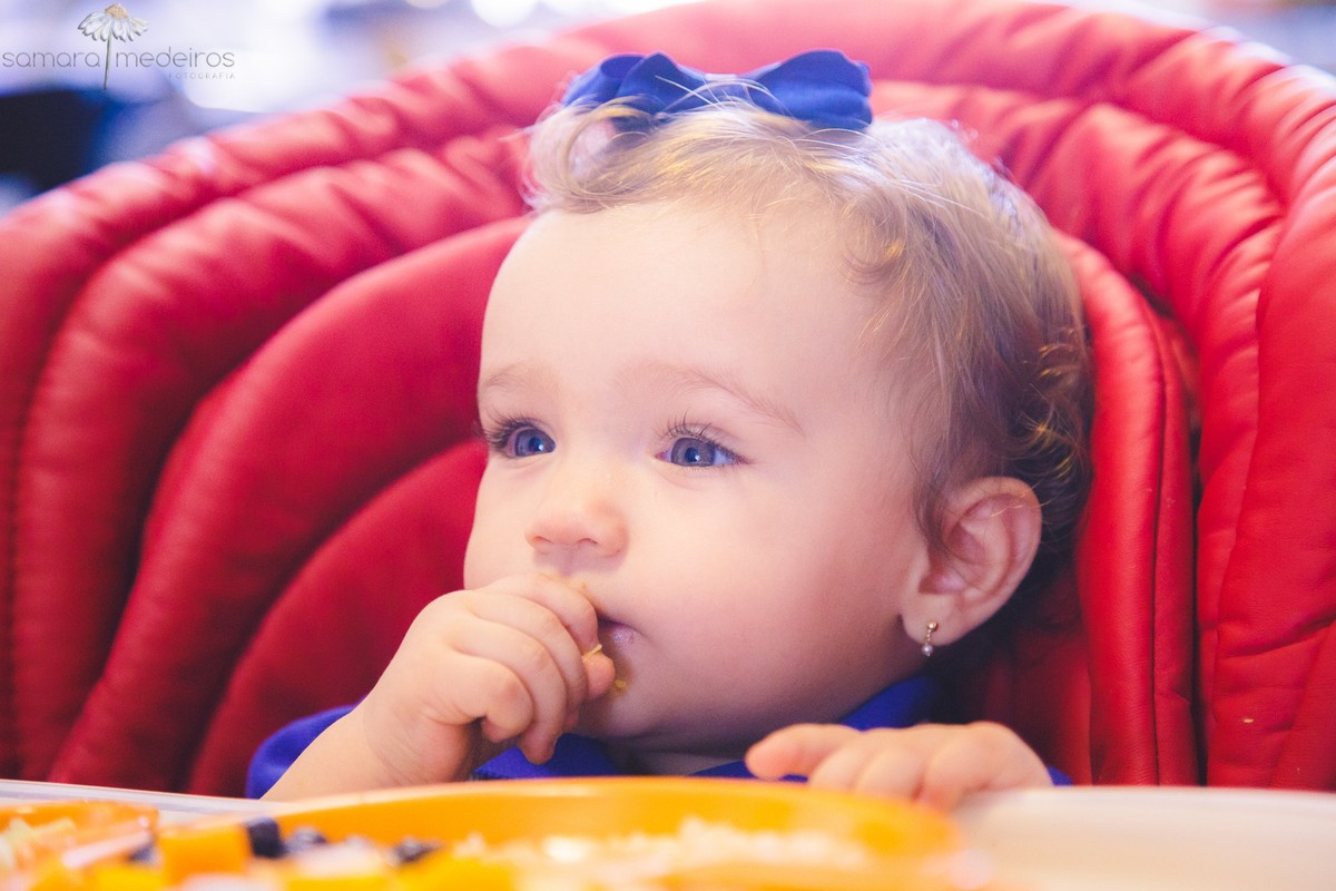 Criança sentada em sua cadeirinha de alimentação comendo o almoço, usando as mãos, durante uma sessão fotográfica infantil.