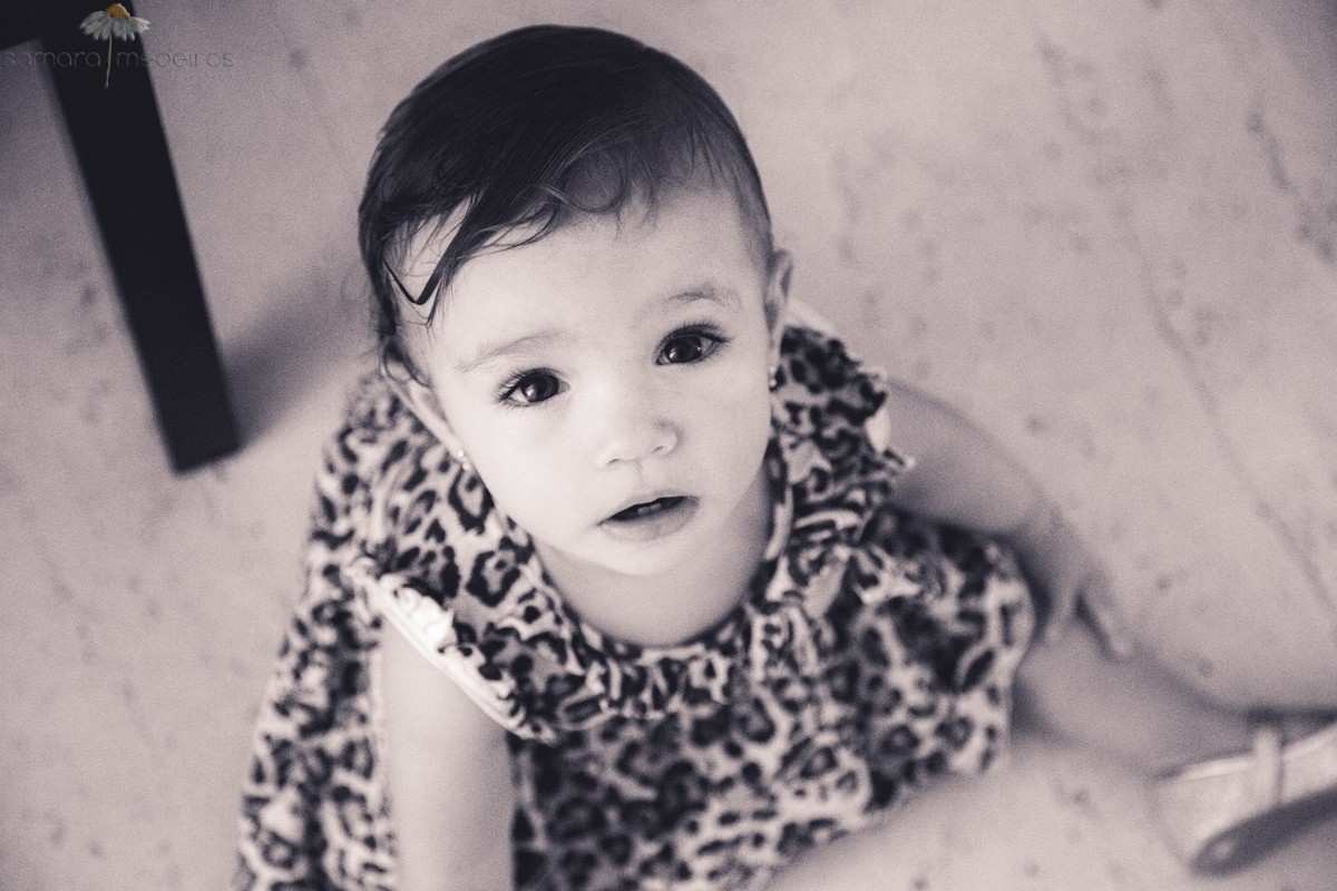 Foto em preto e branco, vista de cima, de uma criança sentada no chão de sua casa e olhando para a câmera. 