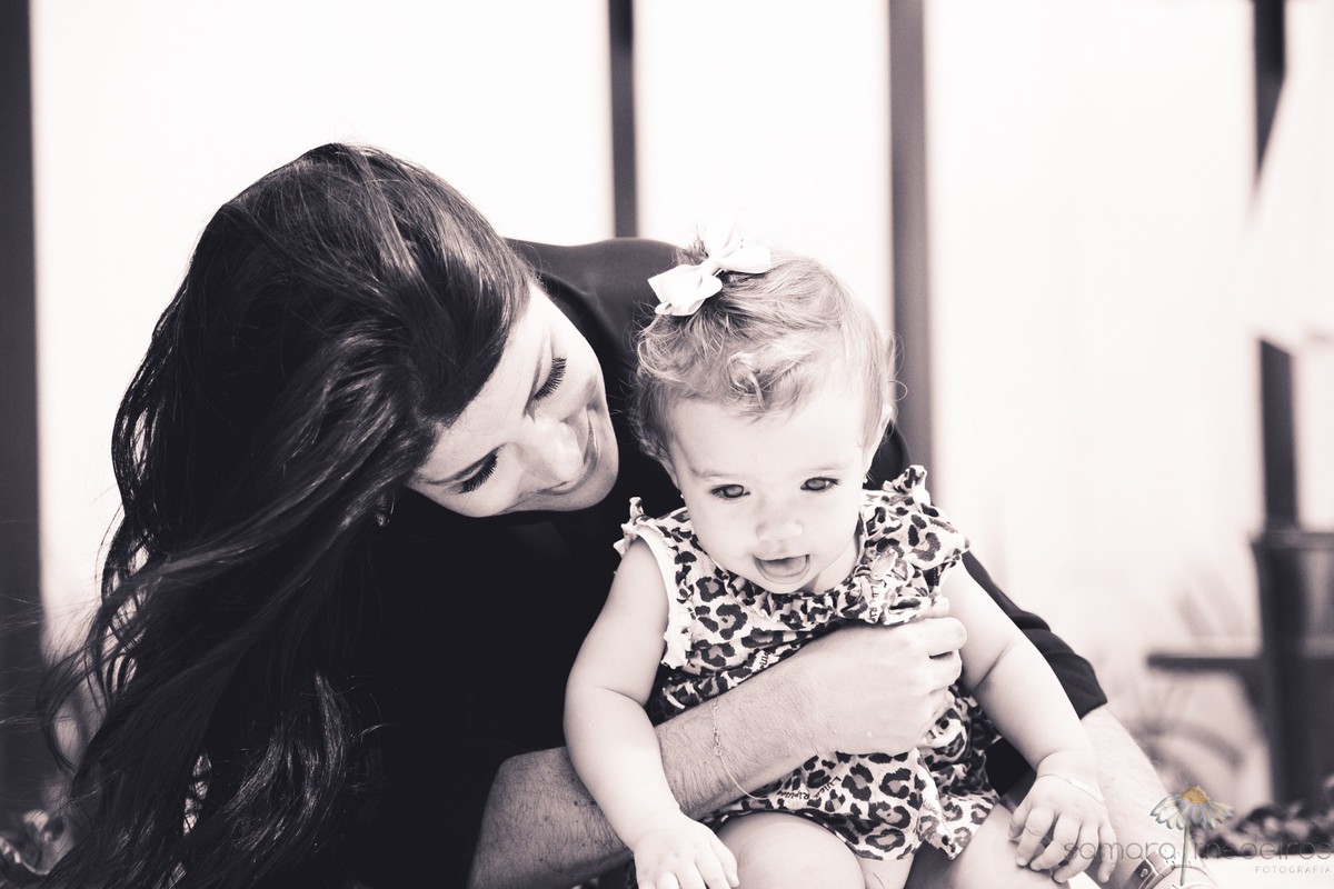 Foto em preto e branco da mãe abraçando a filha e olhando para ela, enquanto brincam na beira da piscina durante ensaio fotográfico lifestyle de família.