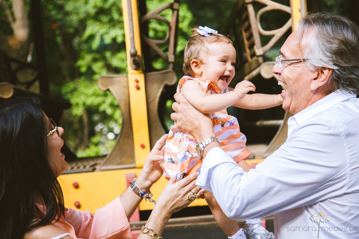 Pai e mãe brincando com a criança de gangorra, enquanto todos os três riem muito e se divertem, durante ensaio fotográfico de família.