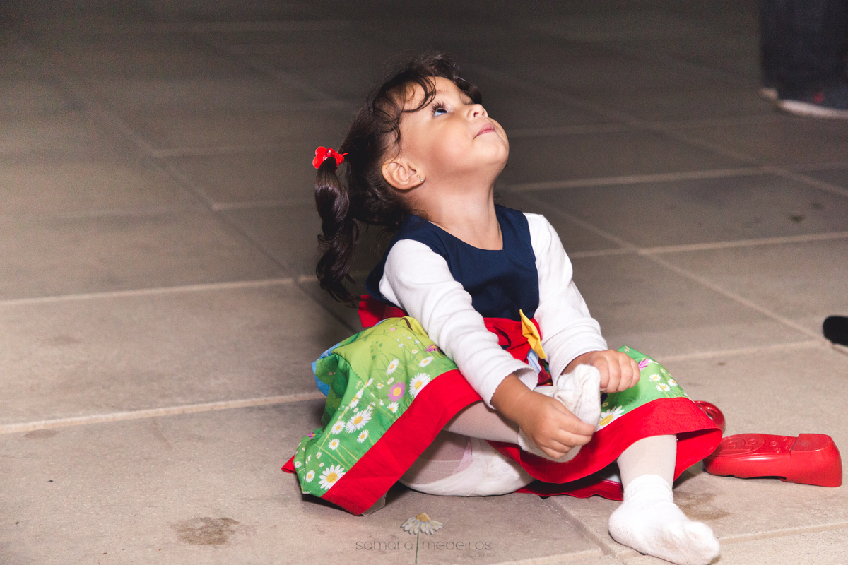Criança sentada no chão, olhando para cima, mexendo no pé para arrumar a meia, durante sua festa de aniversário de 2 anos em Belo Horizonte.