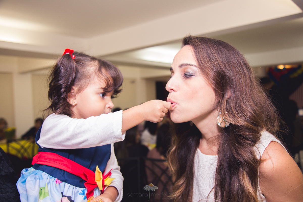 Criança dando gelatina em uma colher na boca da mãe, durante sua festa de aniversário no salão do prédio.