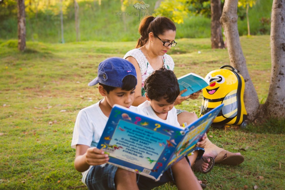 Mãe e seus dois filhos lendos juntos à sombra de uma árvore em um parque de Belo Horizonte.