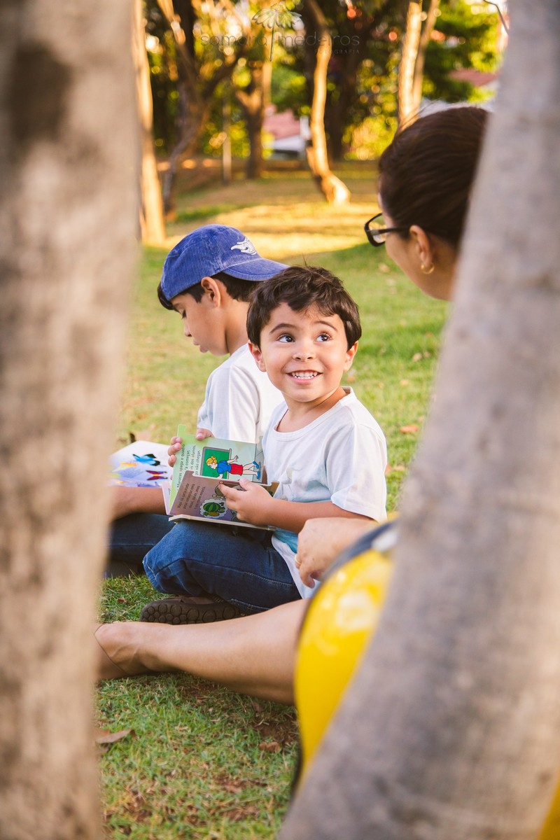 Mãe com seus dois filhos sentados na grama lendo, emoldurados pelos galhos de uma árvore.