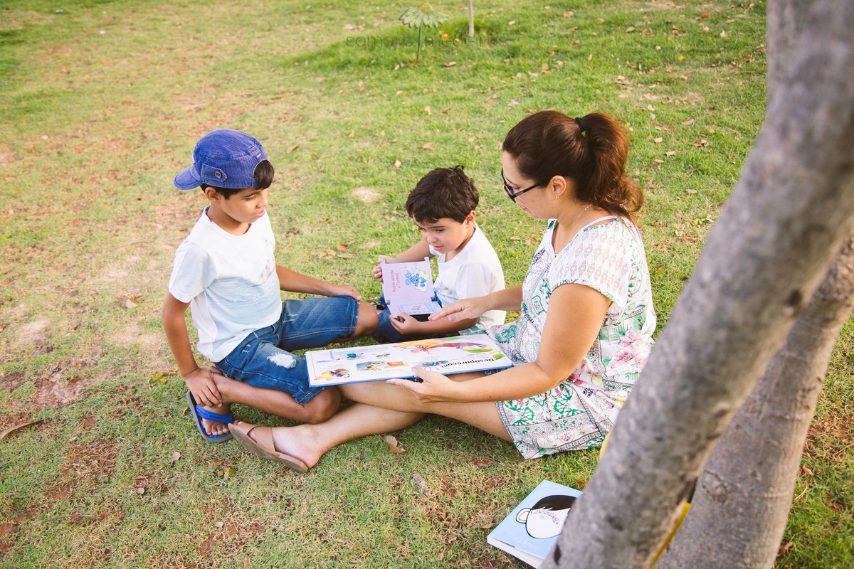 Mãe e dois filhos crianças sentados na grama lendo à sombra de uma árvore, vistos de cima.
