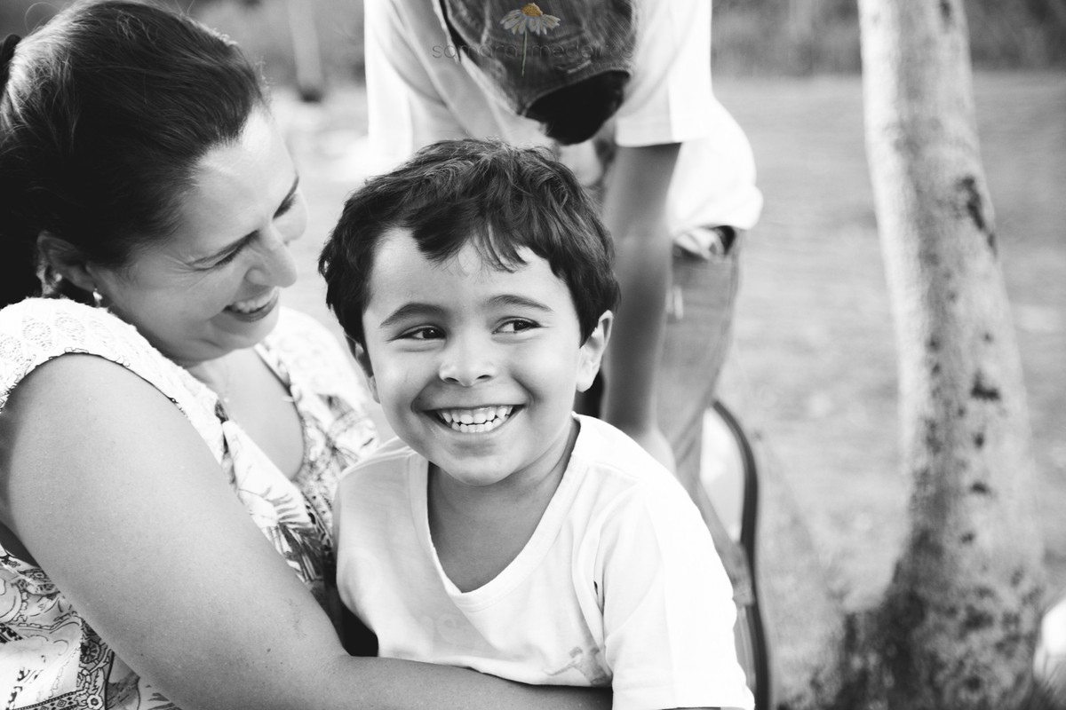 Foto em preto e branco de uma mãe e seu filho sentado em seu colo e ambos sorrindo, durante uma sessão fotográfica de família.