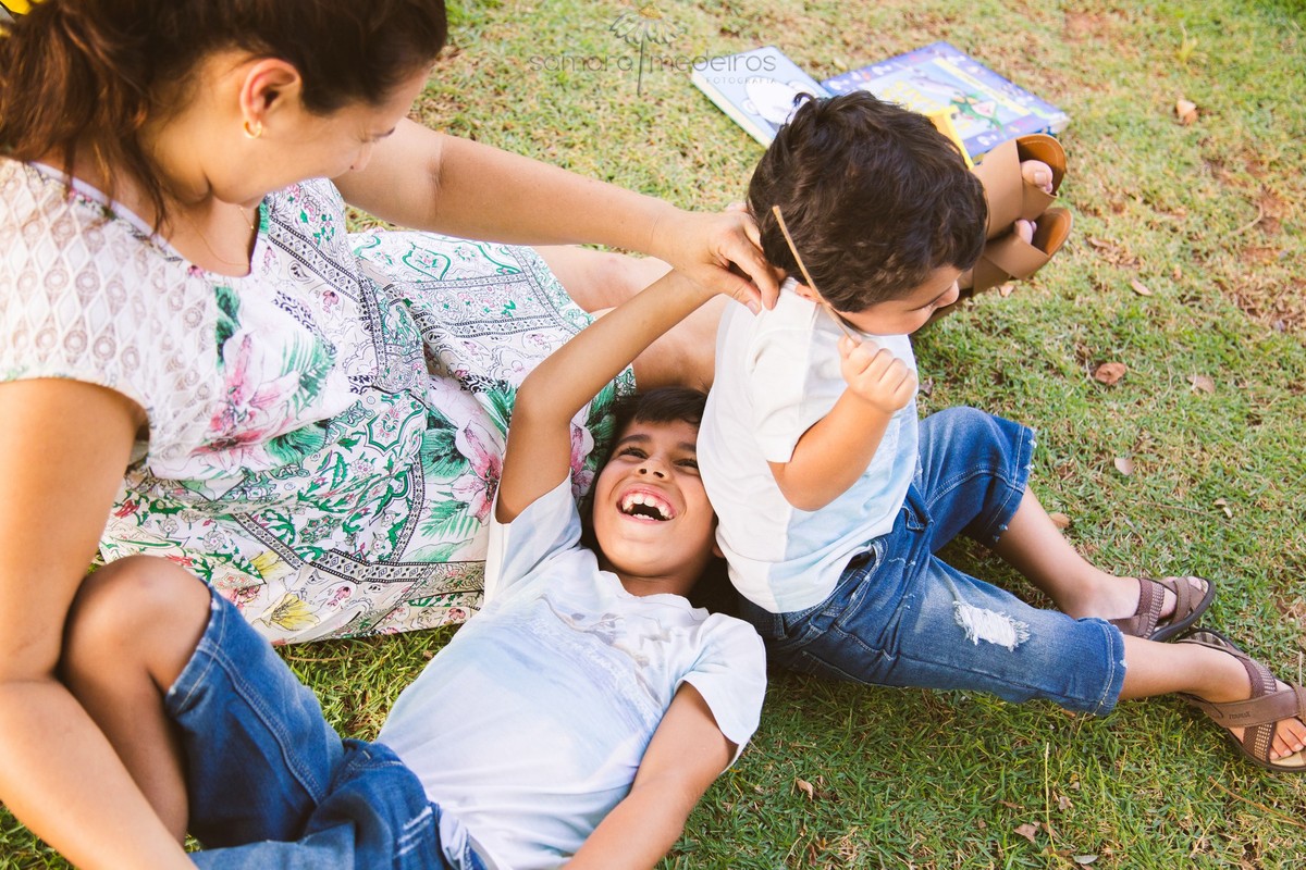 Vista de cima de uma mãe brincando com seus dois filhos, fazendo cócegas neles, todos sentados em um local gramado.