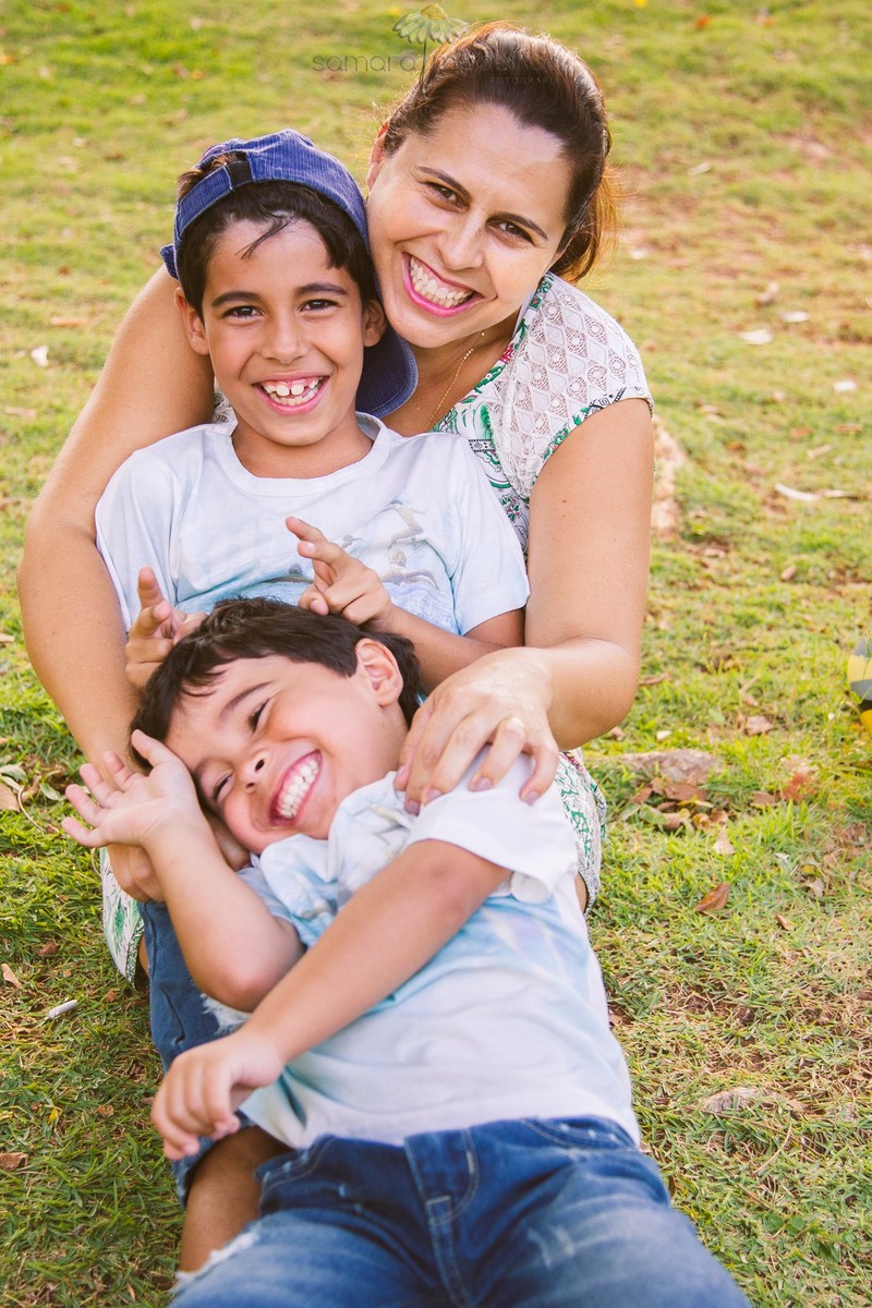 Mãe com seus dois filhos sentados em colo fazendo gracinhas uns com os outros, cócegas, chifrinho para sair na foto, todos descontraídos.
