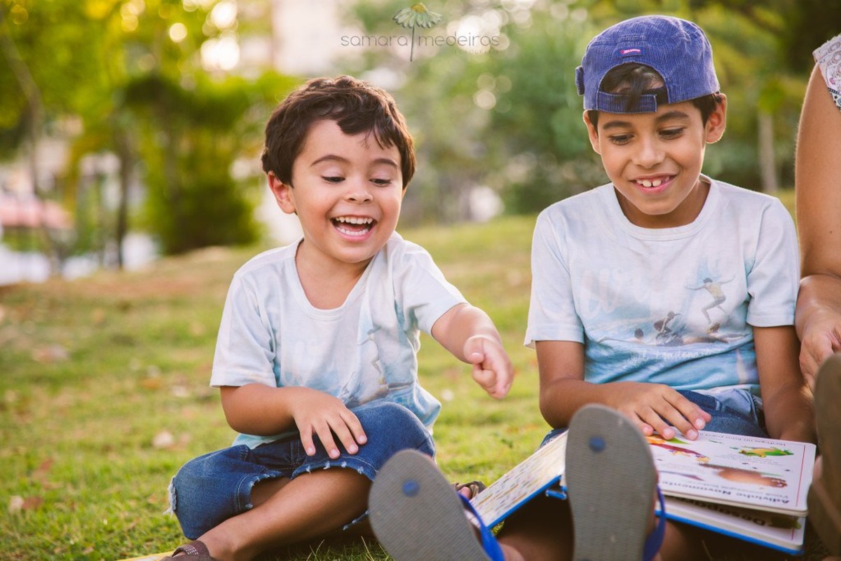 Duas crianças sentadas na grama de um parque com um livro no colo, se divertindo com alguma gravura que viram, rindo e apontando.