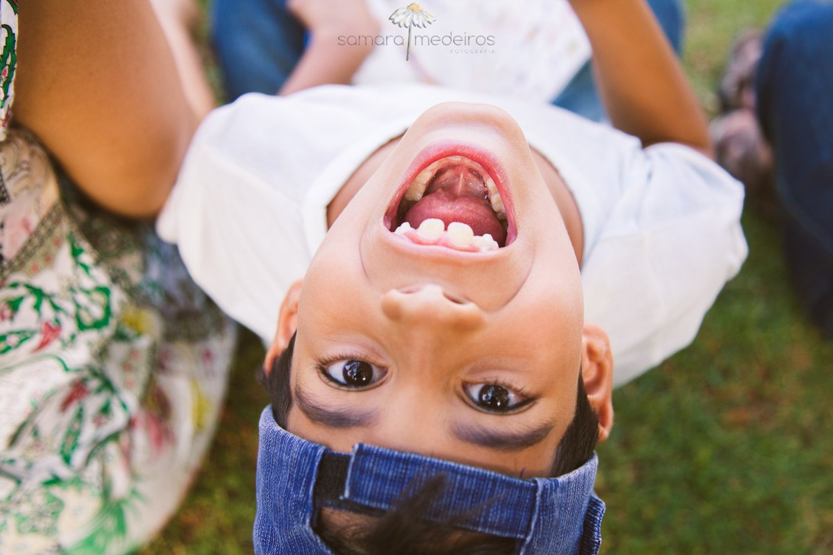 Criança sentada na grama, com o rosto virado para cima, para a foto, fazendo careta, sorrindo, durante uma sessão fotográfica de familia, em Belo Horizonte.