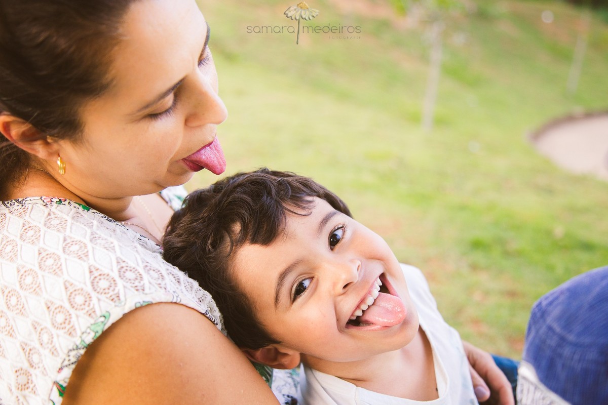 Mãe com seu filho no colo, sentados na grama, ambos fazendo careta com a língua para fora, durante um ensaio de fotos.