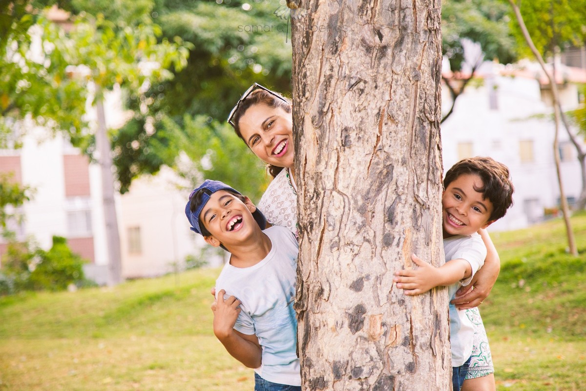 Mãe e seus dois filhos atrás de uma árvore brincando e posando para uma foto durante ensaio de família.