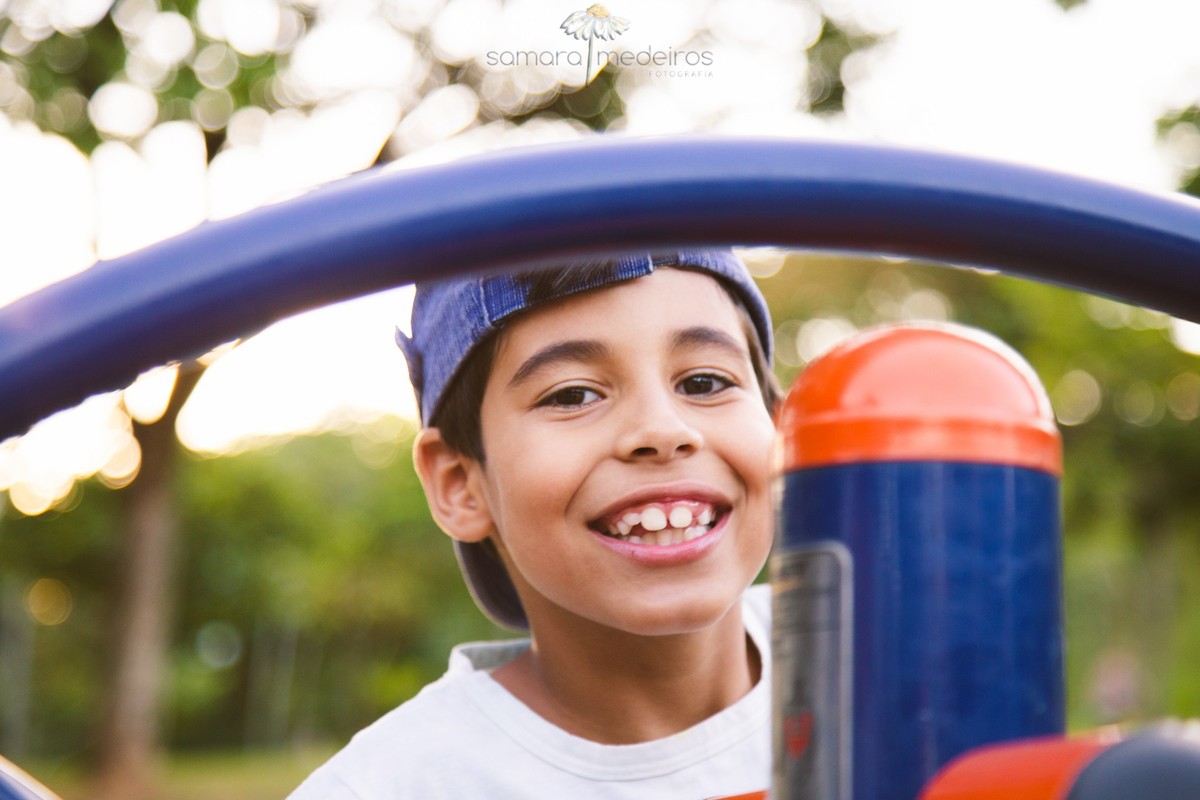 Criança olhando para a foto e sorrindo com o rosto emoldurado por uma estrutura de metal, durante ensaio externo em um parque de BH.