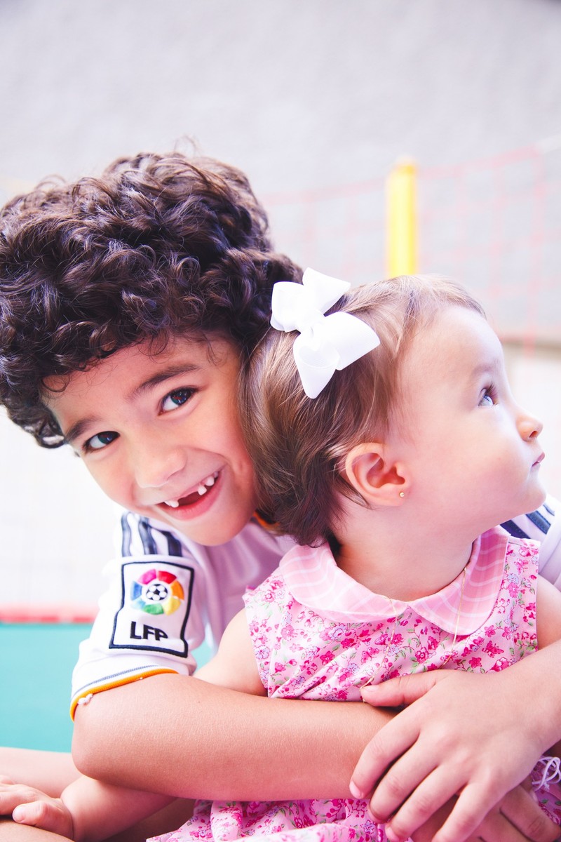 Duas crianças abraçadas, uma olhando para  a câmera e sorrindo enquanto a outra olha para o lado.