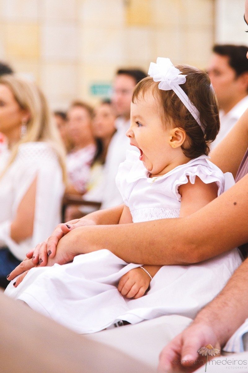 Criança bocejando, sentada no colo da mãe enquanto o padre fala em seu batizado.
